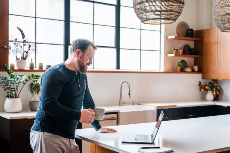A bearded man in dark blue henley stands in modern kitchen viewing laptop on white counter while holding a mug. Bright space with grid windows, wooden cabinets, and plants.