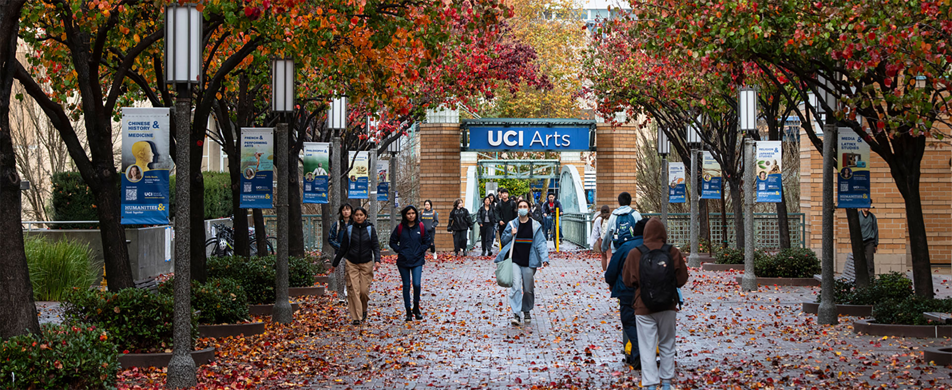 View of a sidewalk with a group of people walking. The sidewalk is covered with fallen, colored leaves.
