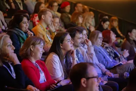 Tiered rows of people sitting and smiling, with one woman leaning forward with her hand under her chin as she listens intently.