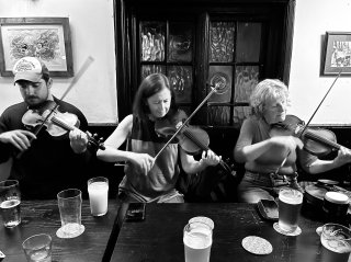 Three fiddlers in a row playing at a pub table; one man and two women.
