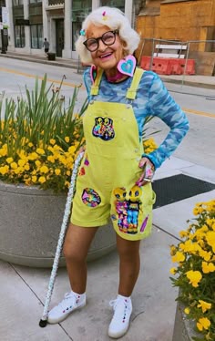 an older woman in yellow overalls and white shoes standing on the sidewalk with a cane