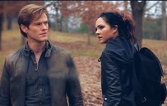 a young man and woman walking through the woods in front of some trees with leaves on them