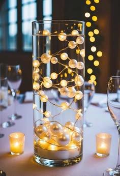 a glass vase filled with white balls and lights on top of a table next to wine glasses