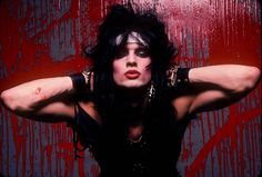 a woman with black hair and makeup holding her hands behind her head in front of a red wall