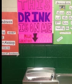 a urinal in a public restroom with posters on the wall behind it that read, this drink is on me