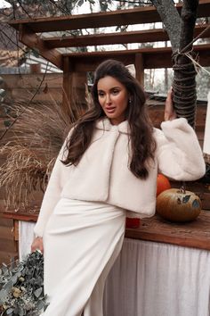 a woman in a white dress and fur coat leaning on a table with pumpkins