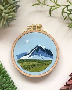 an embroidery project with mountains and trees on the front, surrounded by plants in the background