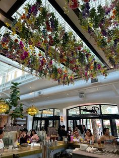 people sitting at tables in a restaurant with flowers hanging from the ceiling and windows above them