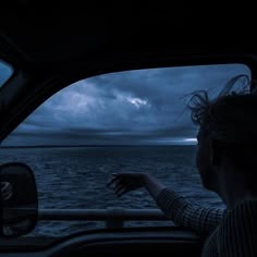 a woman driving a car at night on the ocean with dark clouds in the sky