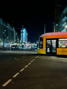 an orange and yellow bus driving down a street at night time with buildings in the background