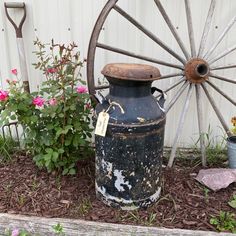an old milk can is sitting in the flower bed next to a wagon wheel and potted plants