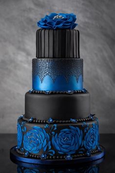a three tiered blue and black cake with flowers on the top is sitting on a table