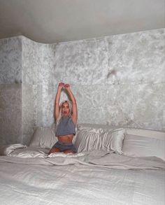 a woman sitting on top of a bed in a room with concrete walls and white sheets