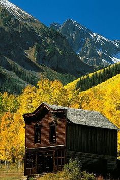 an old wooden building in the mountains surrounded by trees with yellow and green leaves on them