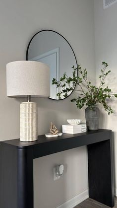 a black table with a mirror and vase on it next to a white plant in a gray room