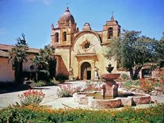 an old building with a fountain in front of it and flowers around the courtyard area