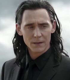 a man with long black hair wearing a suit and tie in front of a cloudy sky