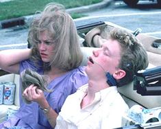 two people sitting in a convertible looking at something on the ground while one woman holds her head