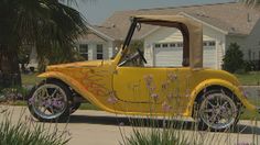 an old yellow car parked in front of a house with palm trees and purple flowers