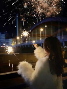 a woman holding a sparkler in her hand and looking at the sky with fireworks
