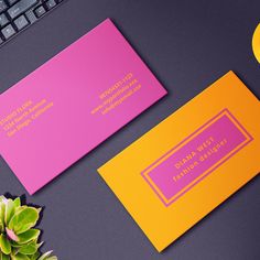 two pink and yellow business cards sitting on top of a computer keyboard next to a plant