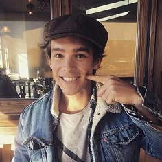 a young man wearing a denim jacket and hat smiles while holding his finger to his nose
