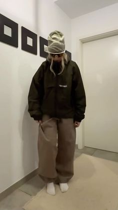 a man standing in front of a white wall wearing brown pants and a black jacket