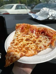 a slice of pizza on a paper plate in someone's hand with cars in the background