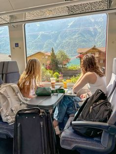 two women sitting at a table with luggage in front of them looking out the window
