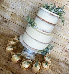 a three tiered cake with flowers and greenery on the top is surrounded by small cupcakes