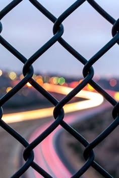 the view from behind a chain link fence looking down at a highway with lights in the distance