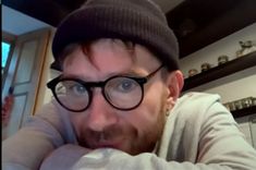 a man wearing glasses and a beanie looks at the camera with his hand on his chin