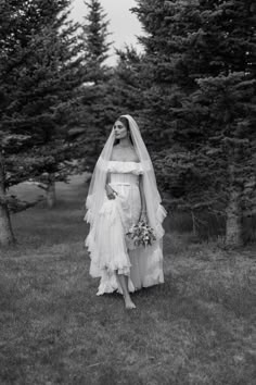 a woman in a wedding dress and veil standing in the grass with trees behind her