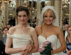 two women standing next to each other in front of a group of people wearing wedding gowns