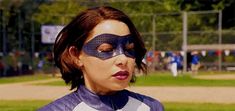 a woman wearing a mask on top of her face at a baseball field with other people in the background