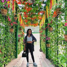 a woman standing in the middle of a walkway covered with plants and hanging baskets filled with flowers