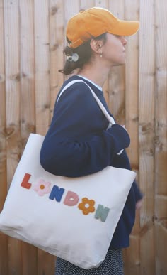 a woman carrying a large white bag with the word london printed on it, in front of a wooden fence