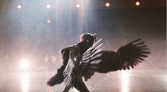 a woman in black is walking with her wings spread out and lights shining behind her