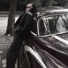 a man leaning on the hood of a car in front of a tree and street