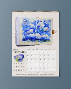 a calendar with watercolors on it and an open book in the middle, sitting on a blue surface