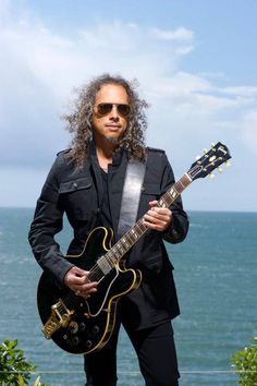 a man with long hair and sunglasses is holding an electric guitar by the ocean in front of him