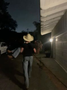 two people walking down the street at night with one person wearing a cowboy hat and holding onto another man's jeans