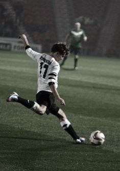 a young man kicking a soccer ball on top of a field with other players in the background
