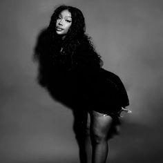 a black and white photo of a woman with long hair wearing a short dress, standing in front of a gray background