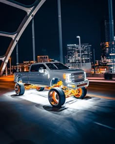 a silver truck with orange wheels driving down the street at night in front of tall buildings
