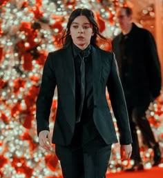 a man in a black suit walking down a red carpeted floor next to a christmas tree
