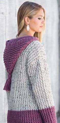 a woman standing in front of a white wall wearing a purple and grey sweater with tassels