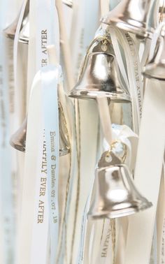 several silver bells hanging from the side of a white wall with ribbon around them and tags attached to it