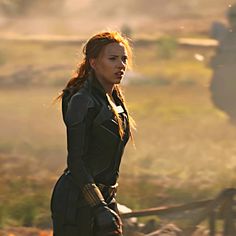 a woman in black outfit standing on grass with fire coming out of her mouth and hands behind her back