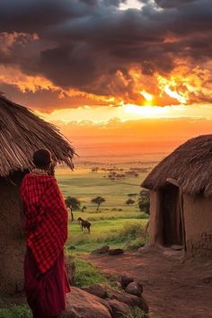 a woman in a red and black dress is looking at the sunset over some huts
